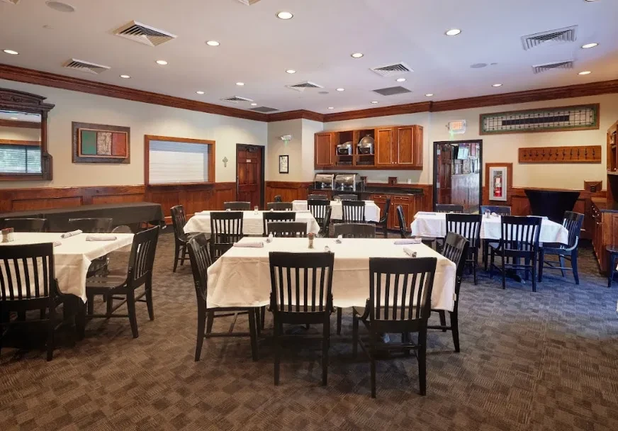 Empty dining room with white-clothed tables, dark chairs, and buffet.