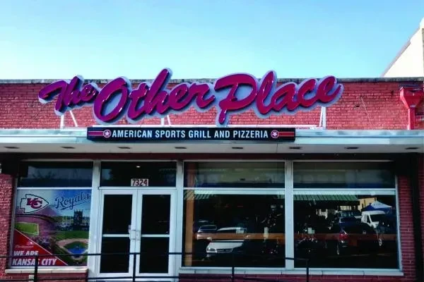 Exterior of "The Other Place" restaurant with neon sign and KC sports decal.
