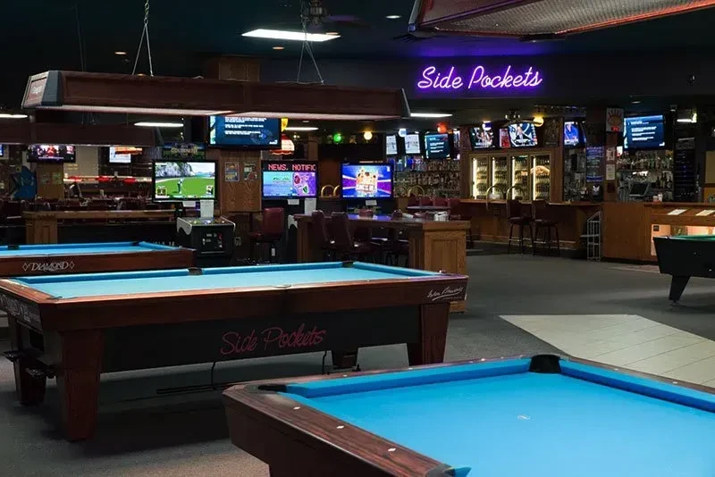 Empty pool hall with blue-felt tables, a bar, and multiple TVs.