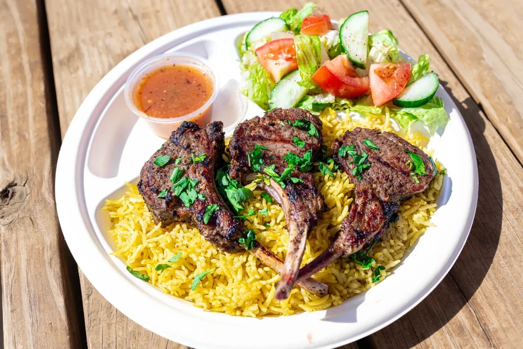 Grilled lamb chops, yellow rice, salad, and sauce on a wooden table.