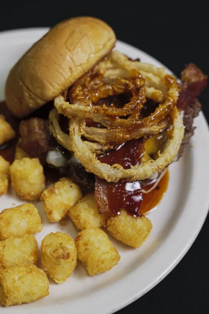Loaded burger with onion rings, dark sauce, cheese, bacon, and tater tots.