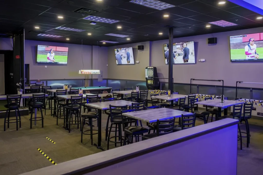 Modern sports bar interior with high-tops, TVs, games, purple ambient lighting.