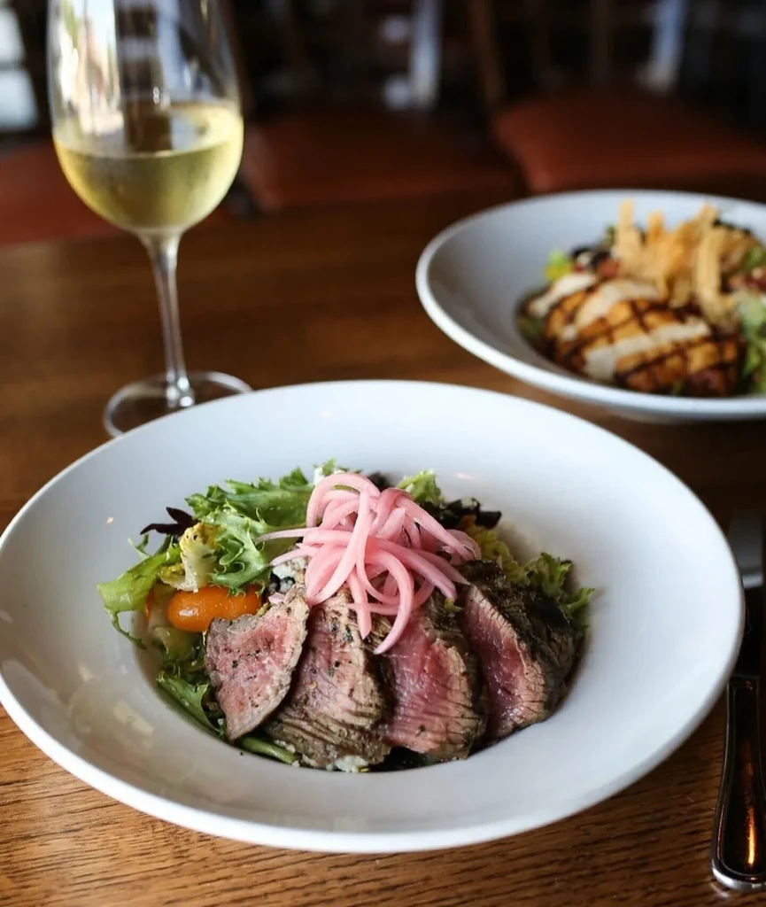 Steak salad, chicken salad, and white wine on a wooden table.