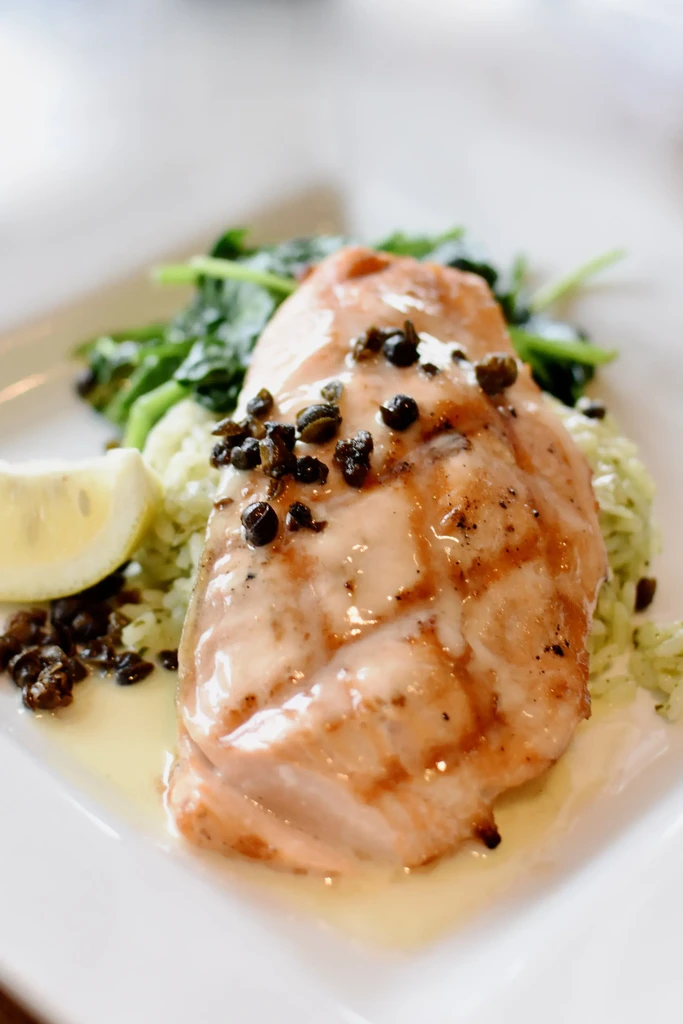 Golden-brown grilled fish with sauce, capers, rice, spinach, and lemon.