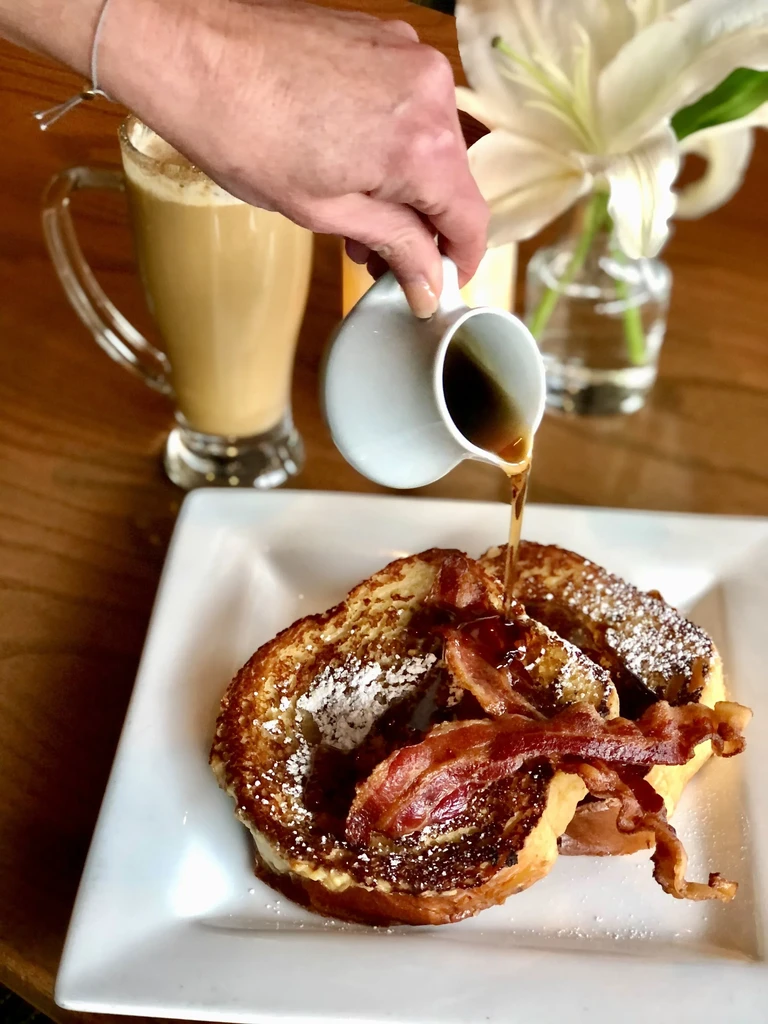 French toast with bacon and powdered sugar, syrup pouring, coffee.