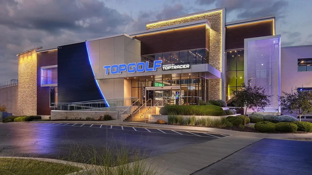 Modern Topgolf building entrance illuminated at dusk, reflected on wet asphalt.
