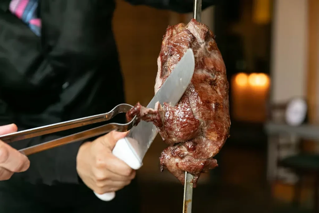 Hands carve browned meat from a skewer; tongs catch a slice.