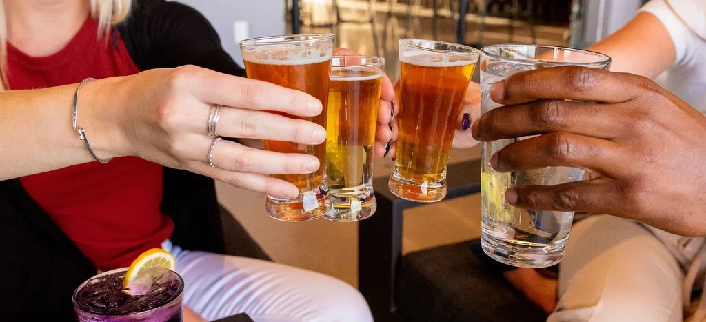 Hands clinking glasses containing beer, clear, and purple drinks.