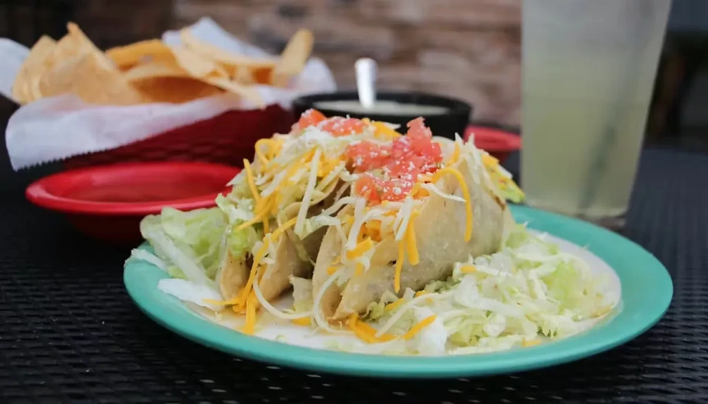 Close-up of three tacos with toppings, tortilla chips, salsa, and a drink.