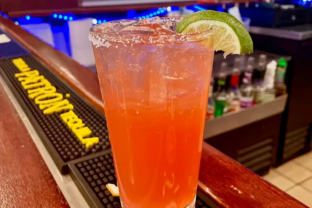 Reddish-orange cocktail with lime, salt rim on dark wooden bar.