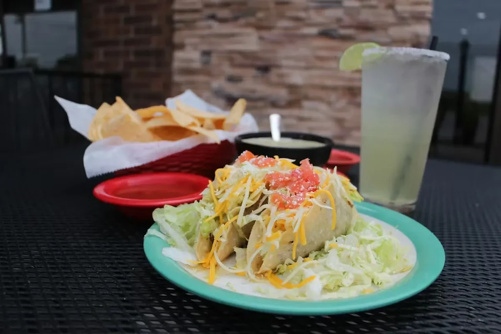 Three hard-shell tacos, chips, salsa, dip, and a margarita outdoors.