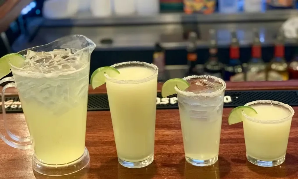 Four lime-green drinks lined on a bar with lime wedges and rims.