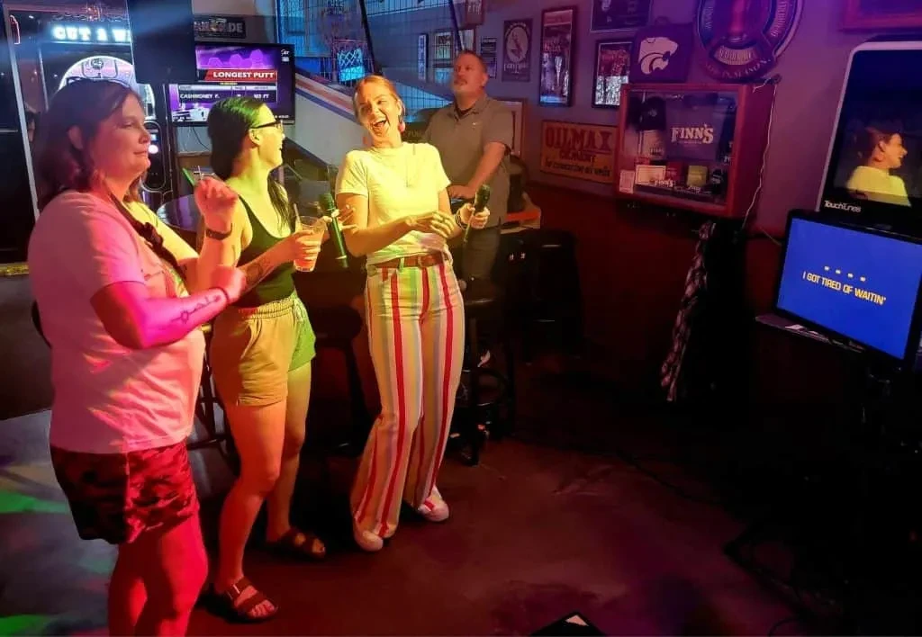 Four people sing karaoke, surrounded by colorful lights and arcade games.