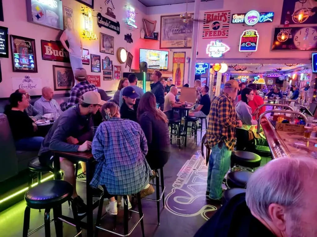 Lively sports bar interior, covered in neon signs, full of patrons.