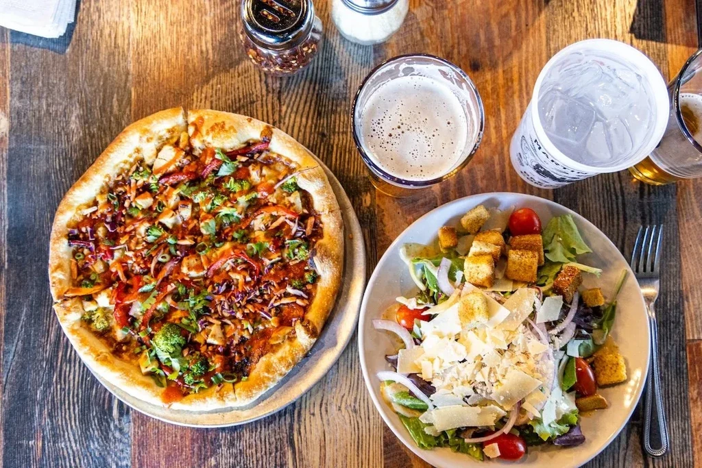 Overhead: Chicken pizza, salad, beer, and water on a rustic table.
