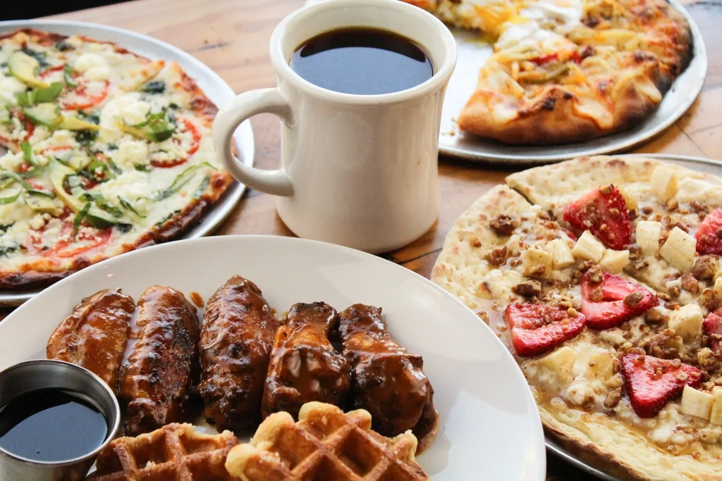 Table of chicken wings, waffles, fruit pizza, savory pizzas, and coffee.