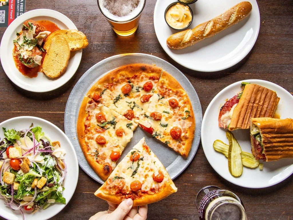Top-down view of pizza (hand holding slice), meatballs, panini, salad, beer.