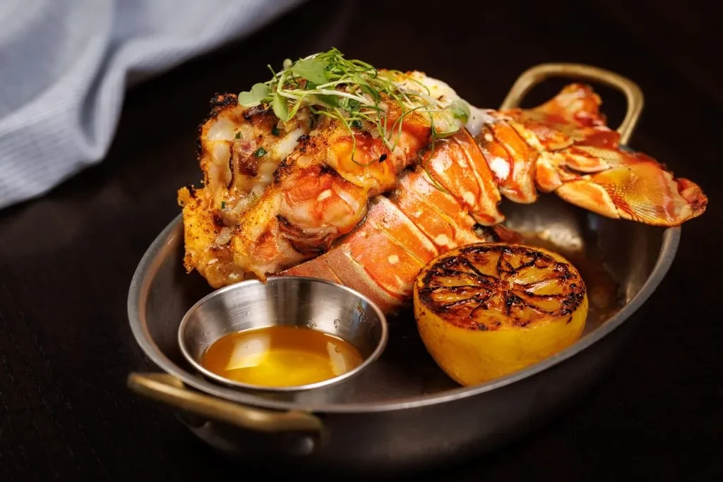 Grilled lobster tail in dark pan with butter, lemon, microgreens.