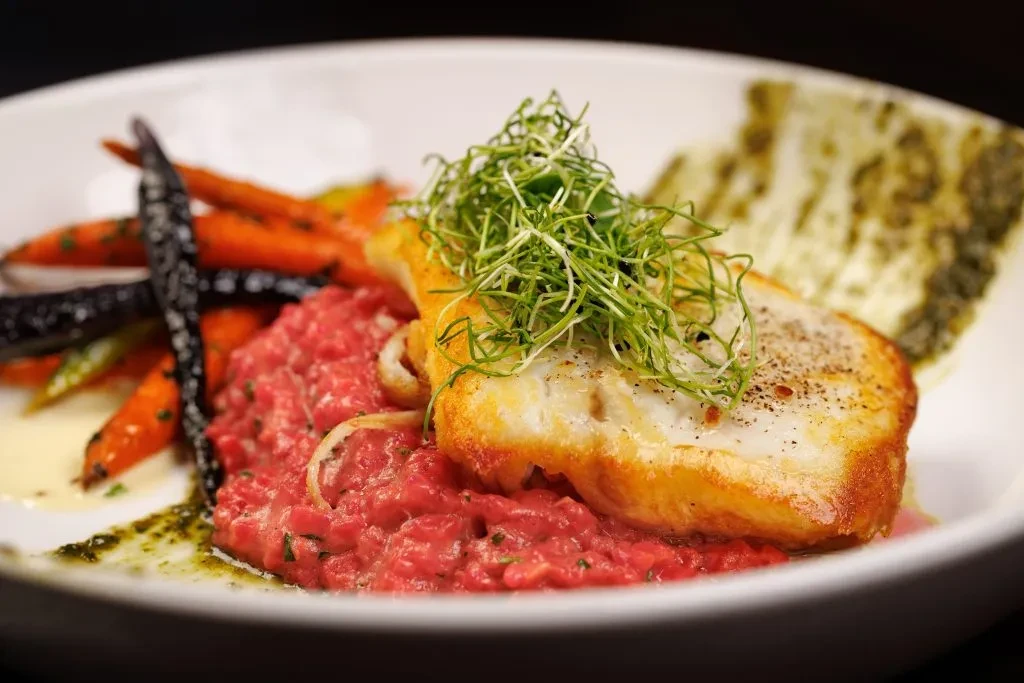Professionally plated pan-seared fish fillet, pink risotto, roasted carrots, microgreens.