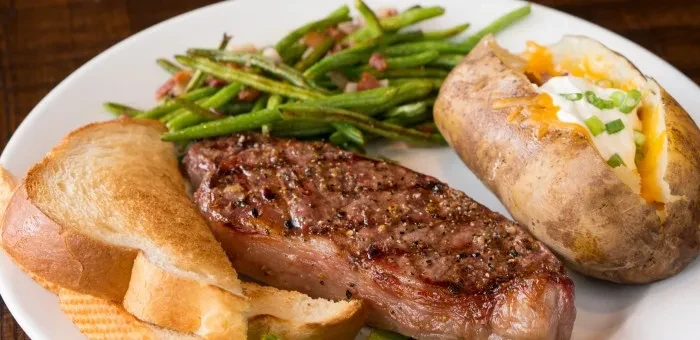 Plated grilled steak, baked potato, green beans, and toast.