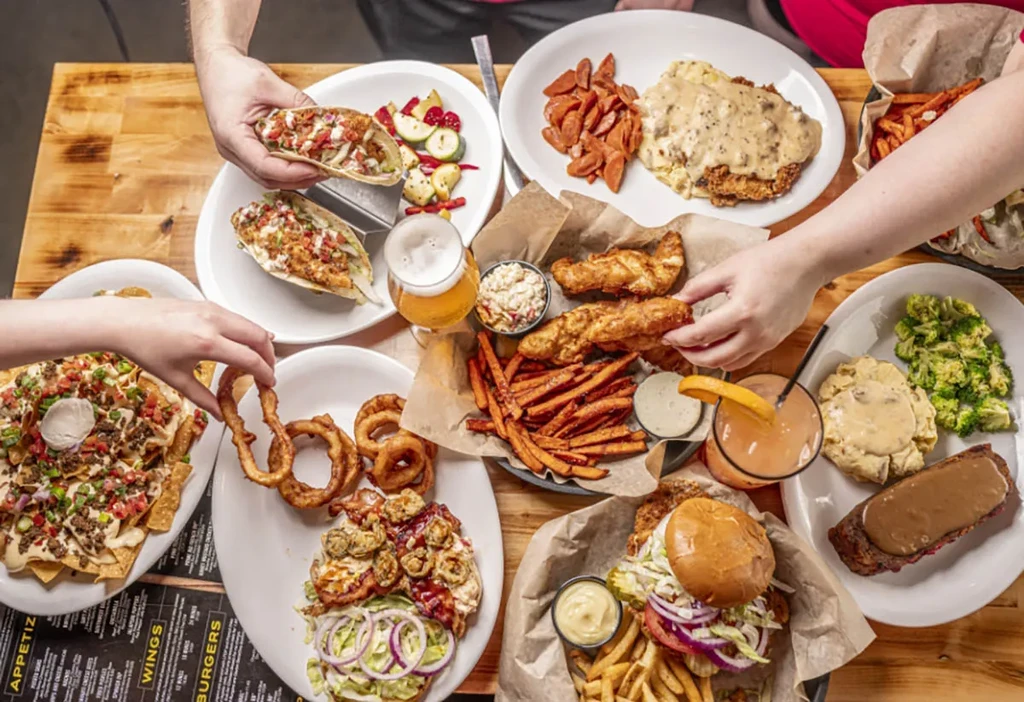 Hands share various American comfort foods and drinks on a wooden table.