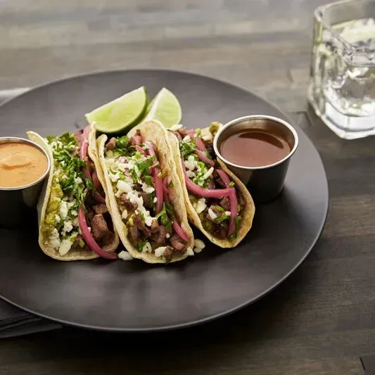 Three tacos with meat, cheese, herbs, pickled onions, limes, and sauces.