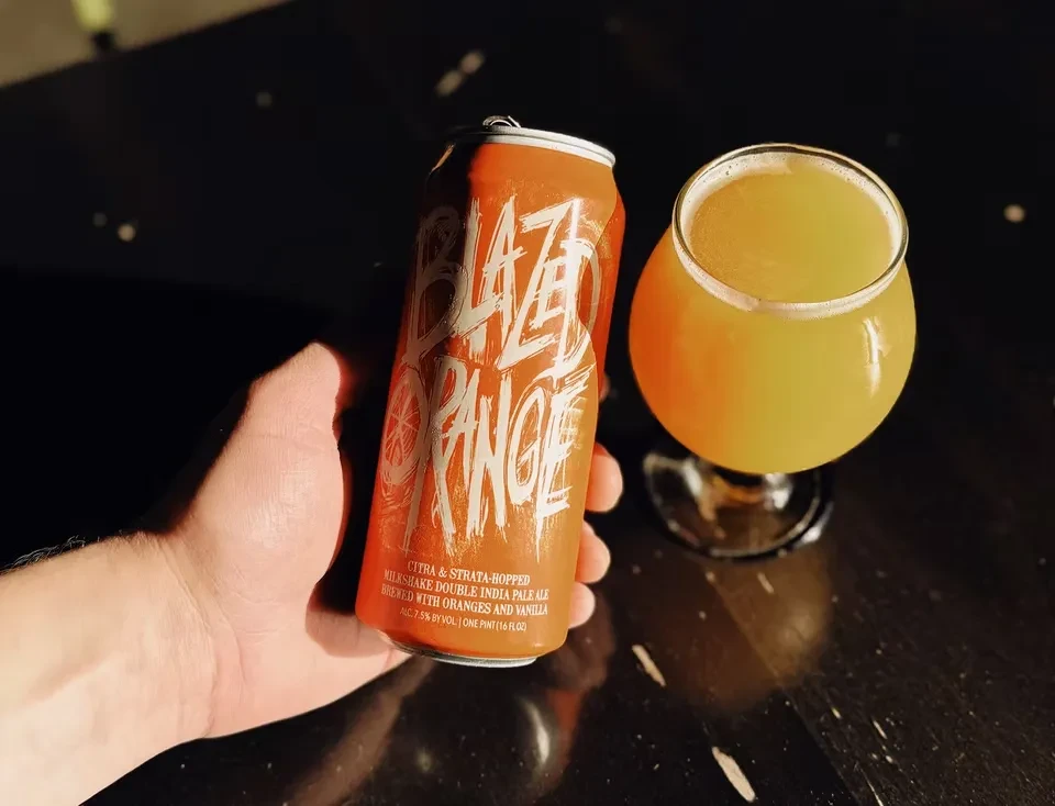 Hand holds "BLAZED ORANGE" beer can and glass of hazy orange ale.