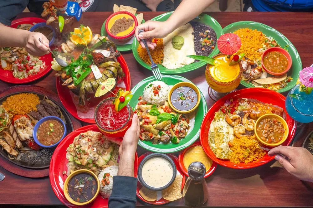 Overhead view: hands sharing a vibrant Mexican food and drink spread.