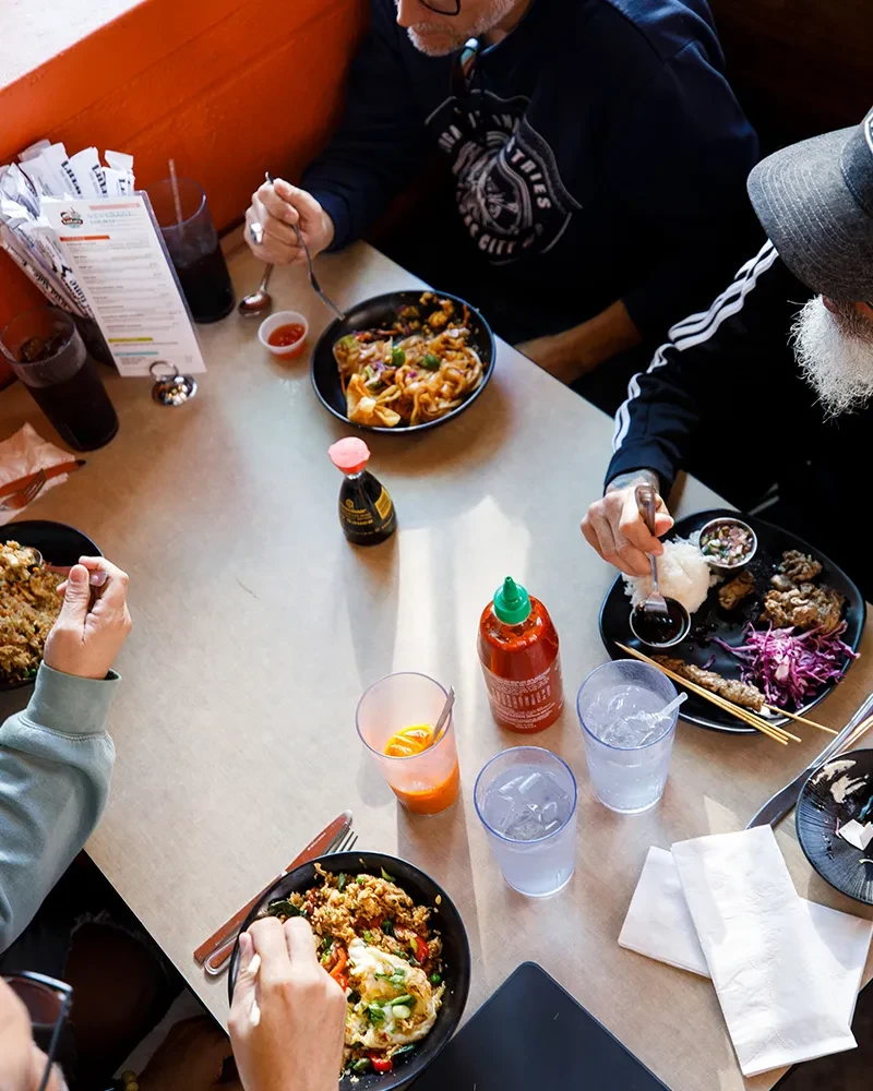 Overhead view of people sharing Asian noodles, fried rice, and skewers.