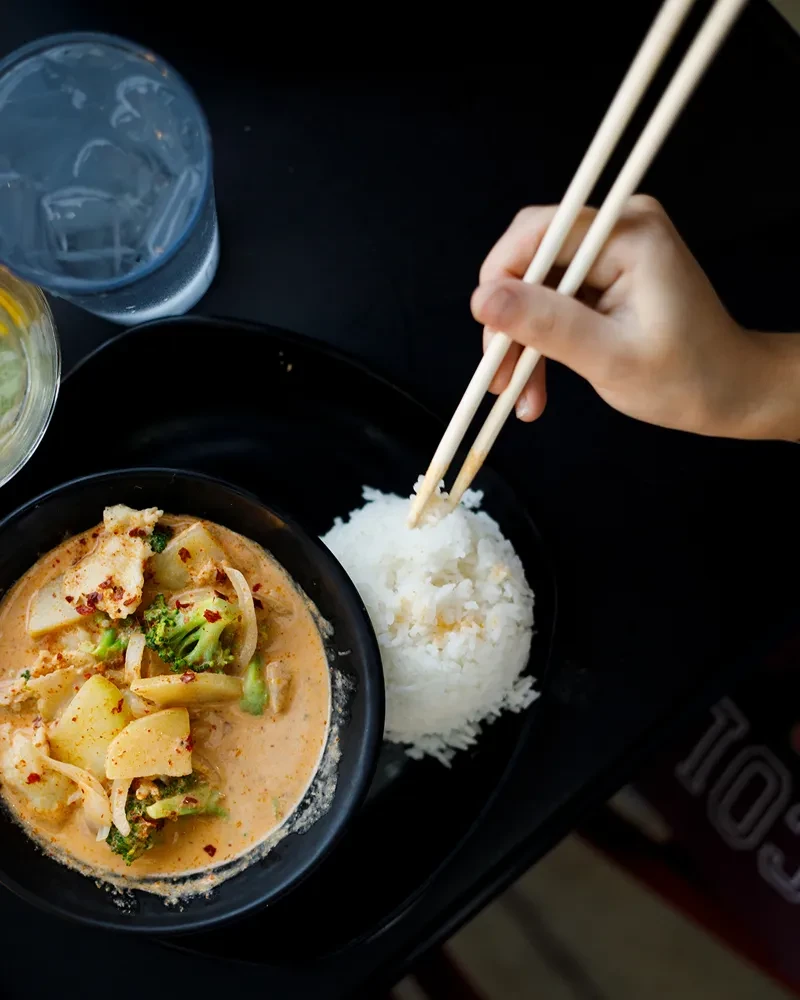 Hand with chopsticks picking rice next to a bowl of curry.