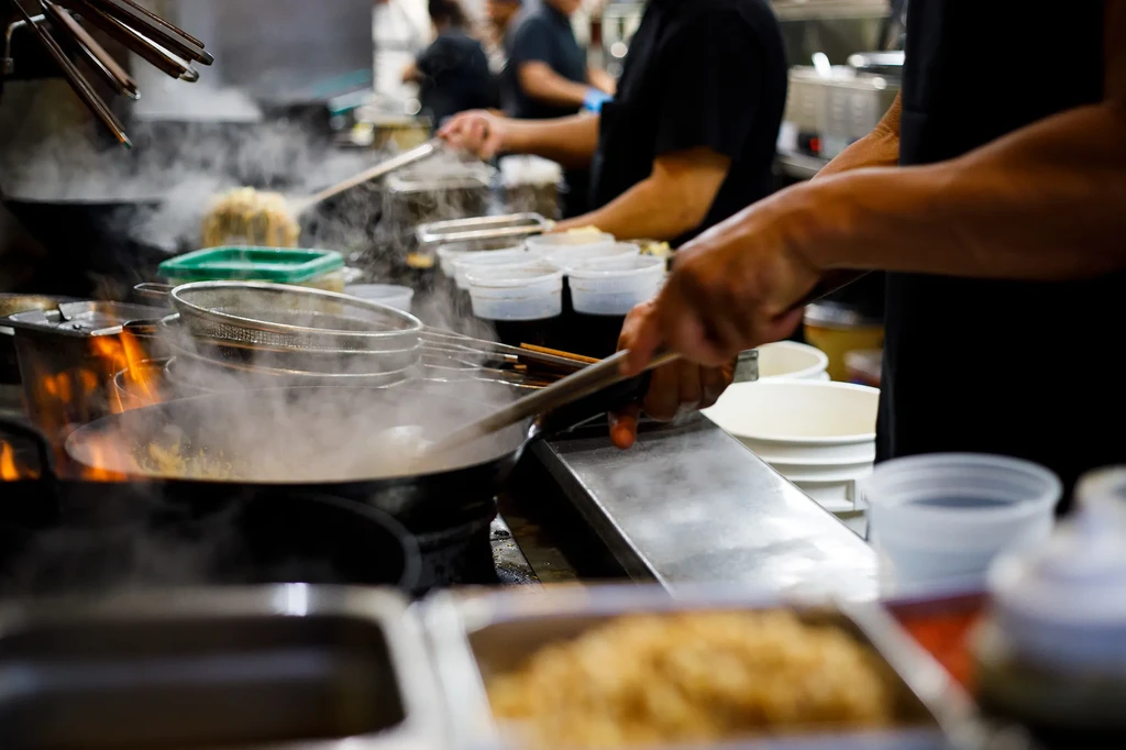 Chef's hands stir food in a flaming wok in a bustling kitchen.