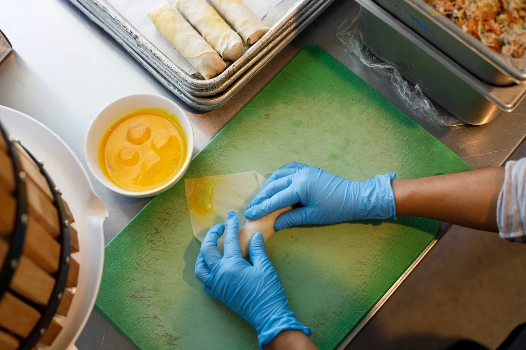 Blue-gloved hands roll spring rolls on a green board, with filling.