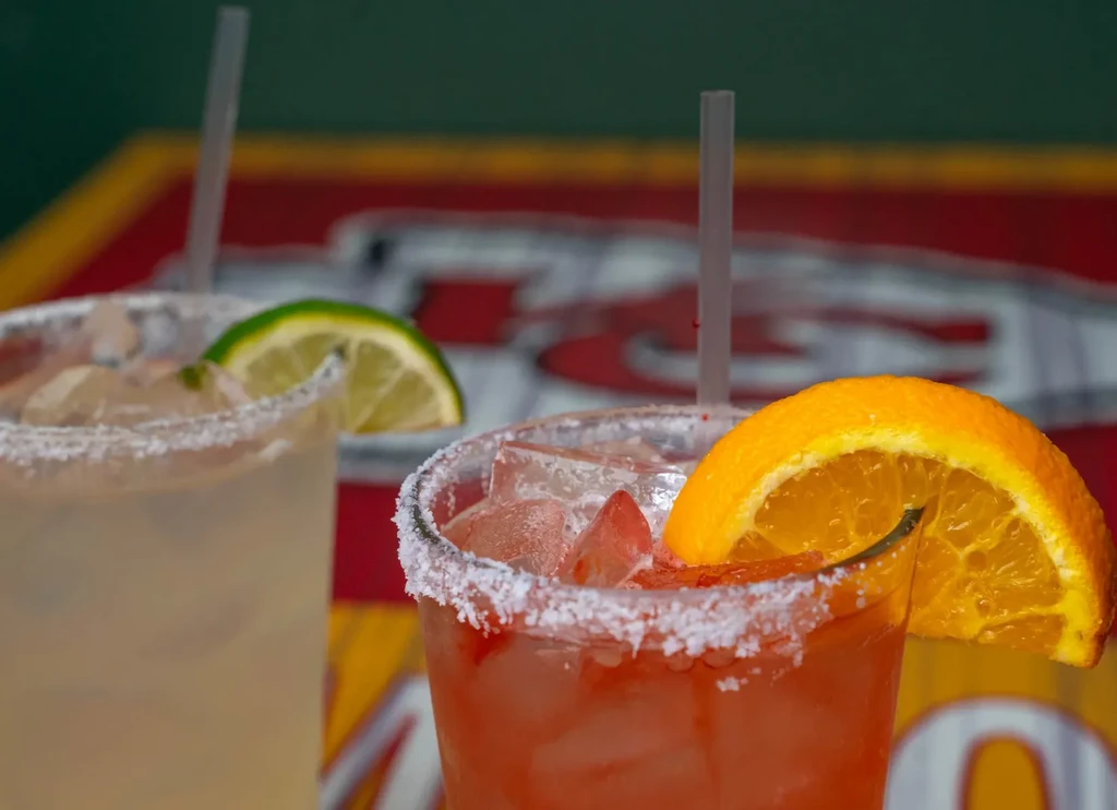 Two colorful garnished cocktails with salt rims and fruit slices.