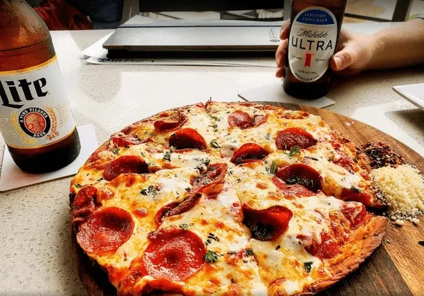 A pepperoni pizza, two beers, and condiments on a wooden board.