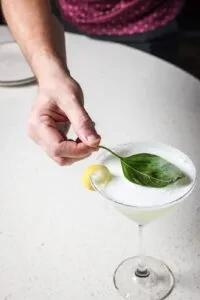 Close-up: Hand garnishing frothy cocktail with basil leaf and lemon peel.