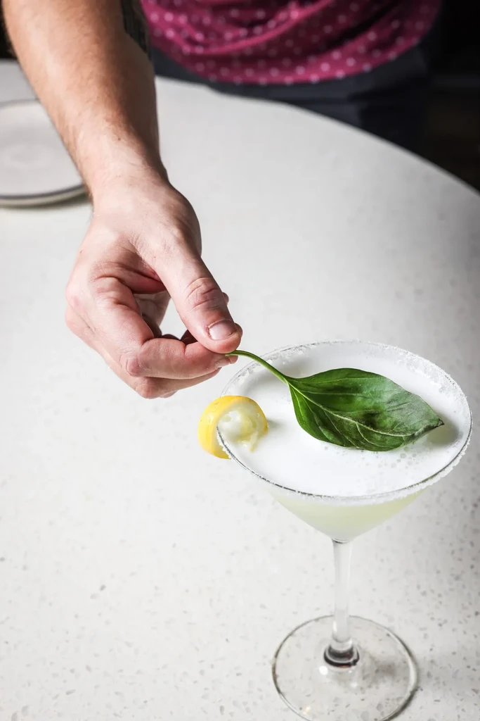 Hand places basil into greenish-yellow cocktail in a martini glass.