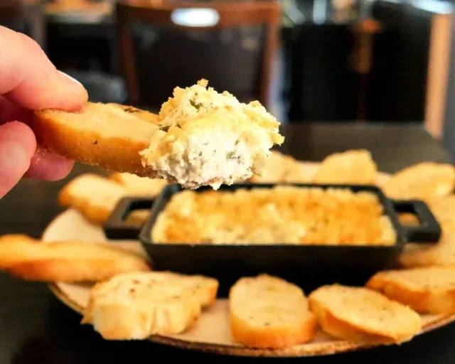 Hand scooping creamy, herb-speckled dip onto toasted bread slices.