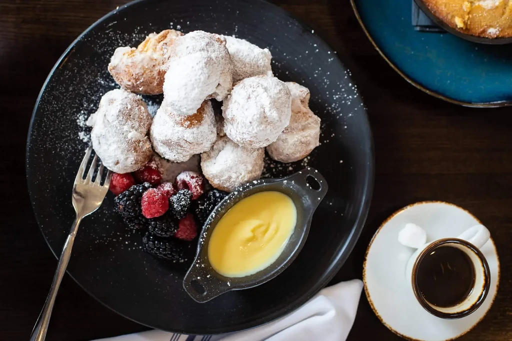 Overhead: Powdered sugar dough balls, berries, sauce, and coffee on a plate.