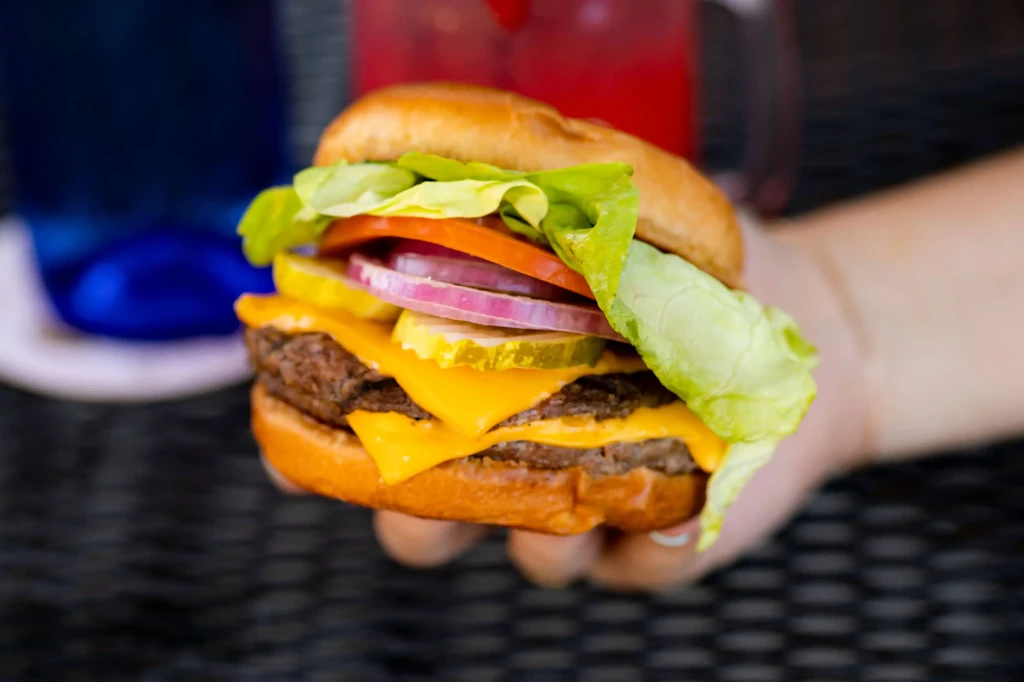 Hand holds a double cheeseburger with cheese, lettuce, tomato, onion, pickles.