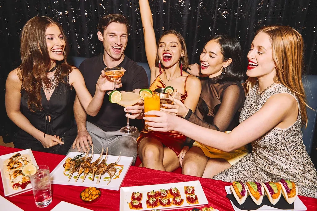 Five smiling young adults toast cocktails at a restaurant table with food.