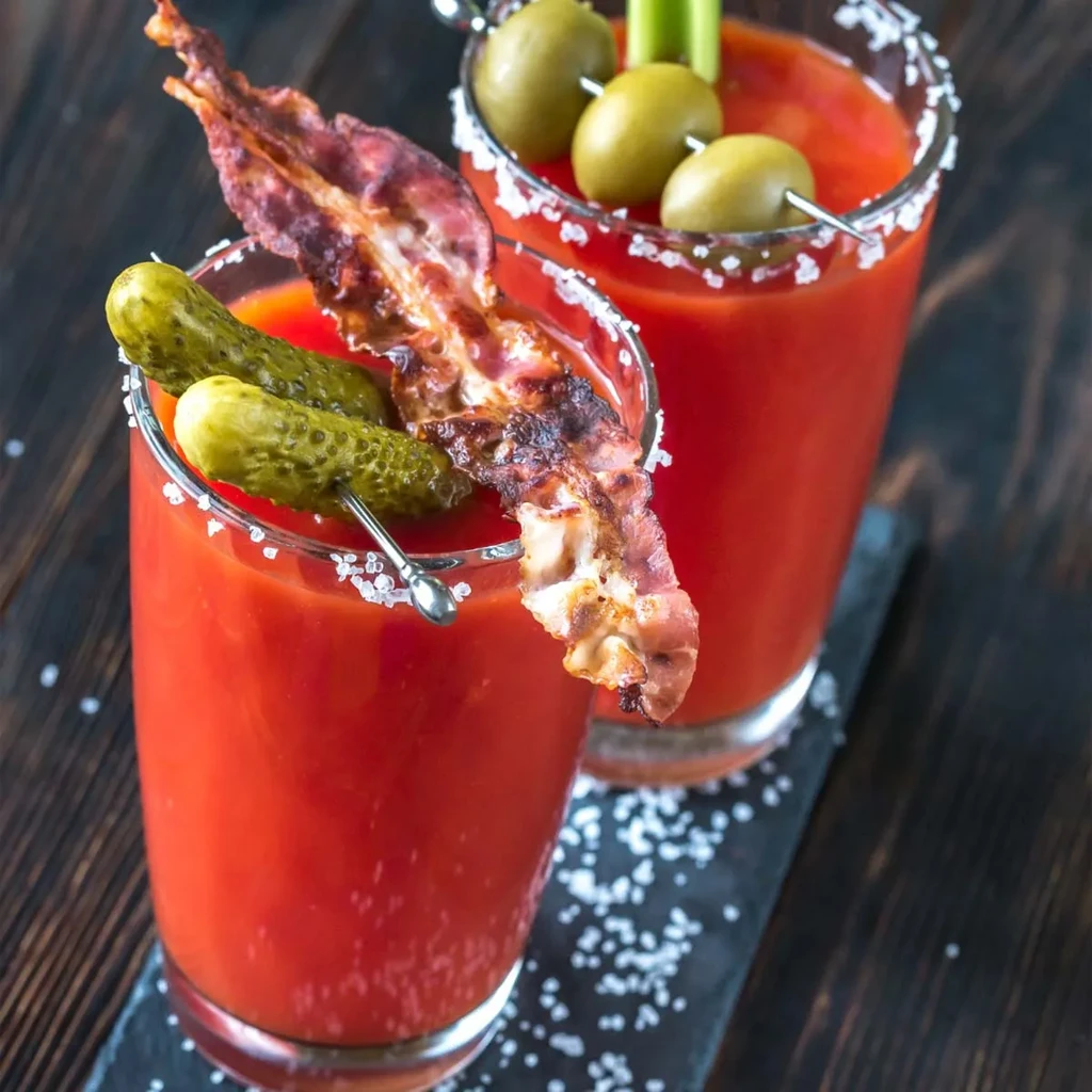 Two Bloody Mary cocktails with elaborate garnishes on a dark surface.