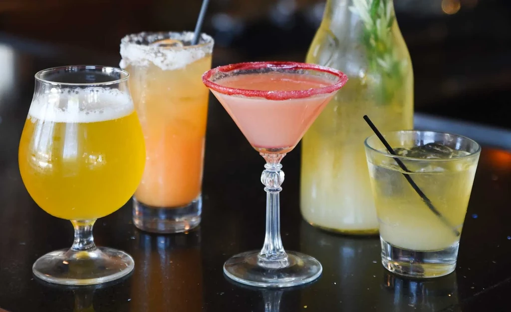Five colorful drinks, including beer and cocktails, on a dark table.