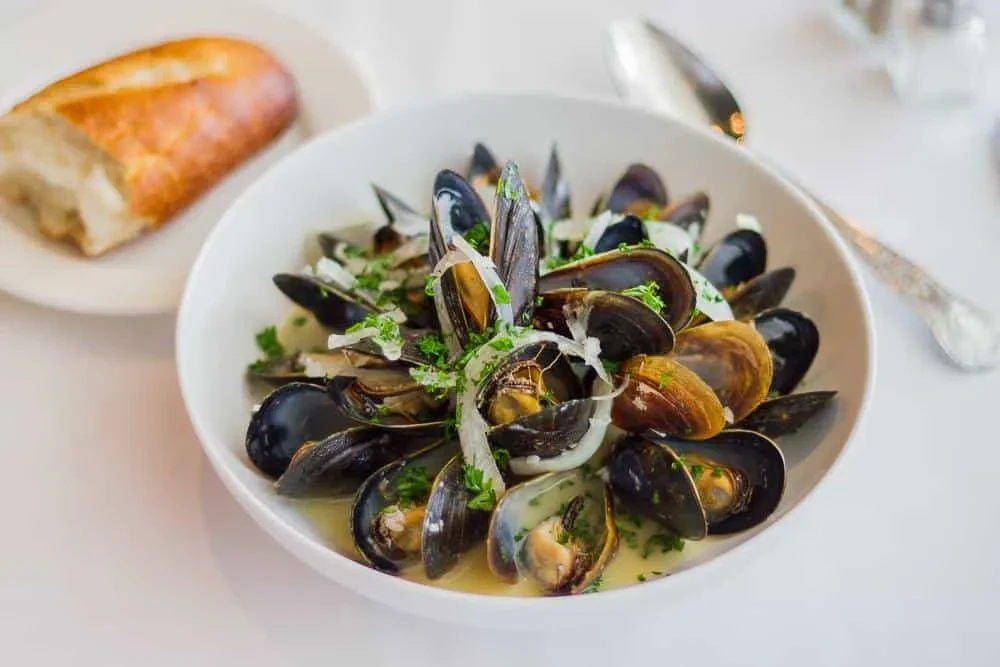 Bowl of mussels in creamy broth, garnished with herbs and bread.