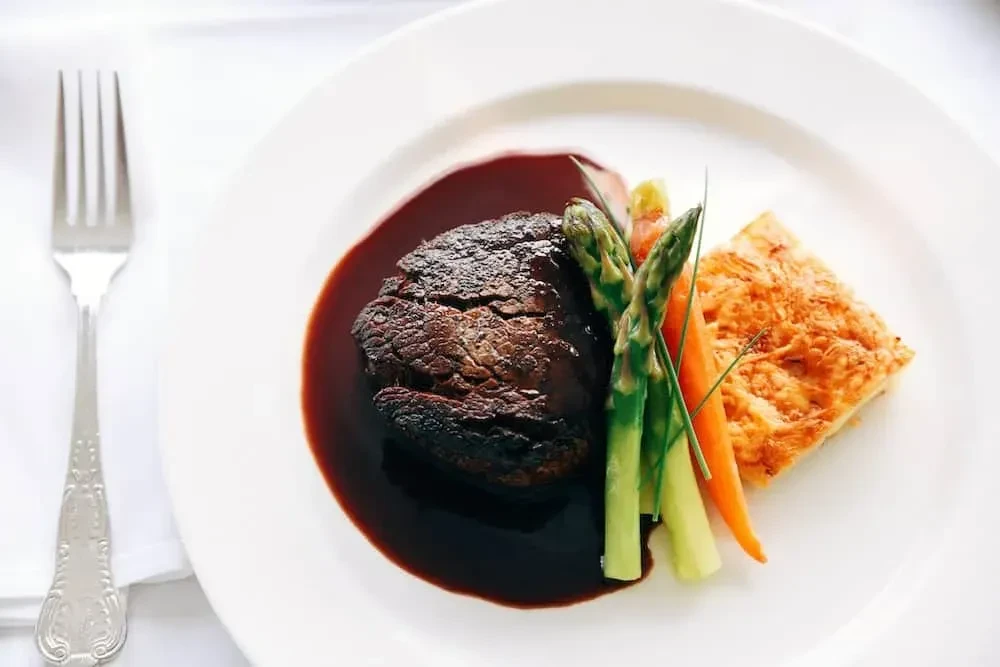 Seared meat with sauce, asparagus, carrots, and gratin on white plate.