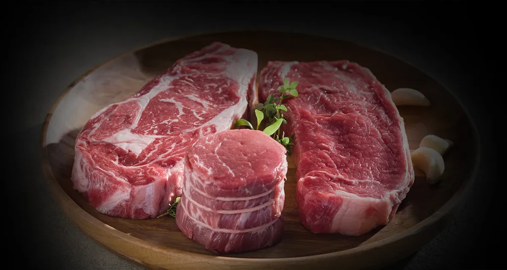Raw ribeye, leaner steak, and tied tenderloin with herbs on wood.