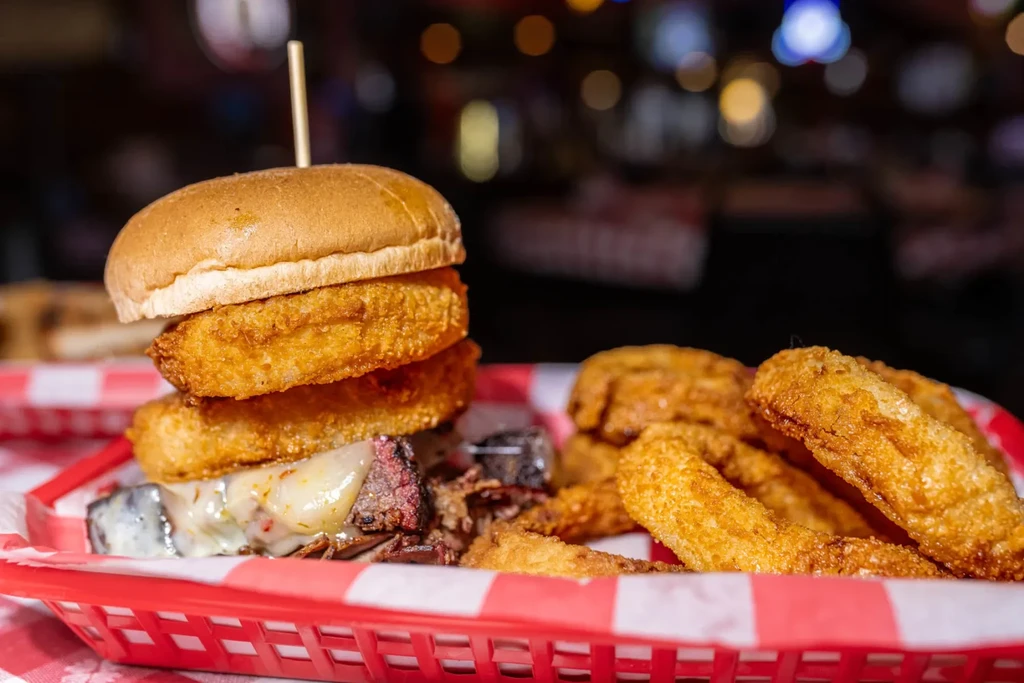 Stacked shredded meat burger, cheese, and onion rings in red basket.
