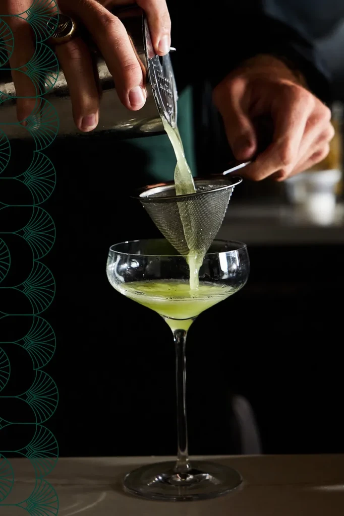 Bartender's hands pour greenish-yellow cocktail into a coupe glass.