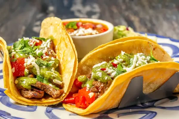 Two tacos in a metal holder, filled with meat, vegetables, and cheese.