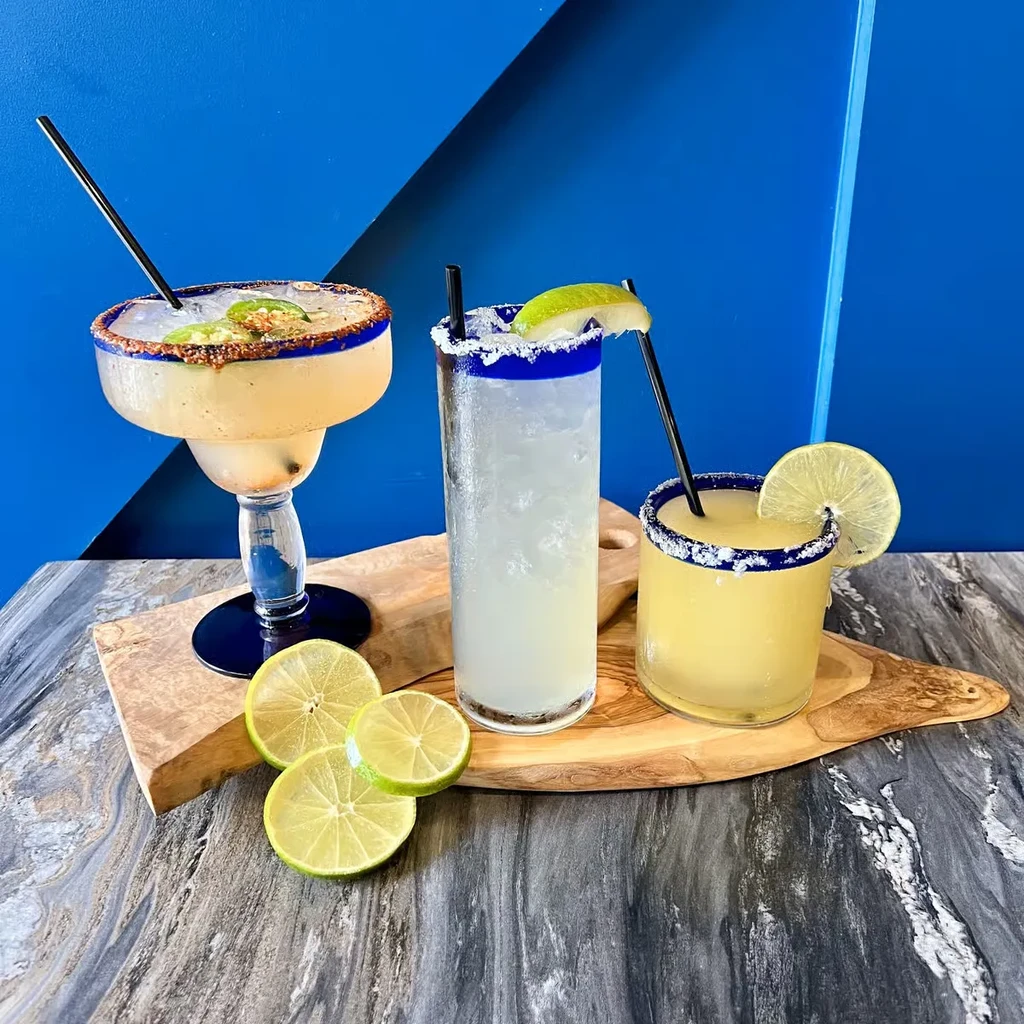 Three diverse cocktails on a wooden board against a blue wall.