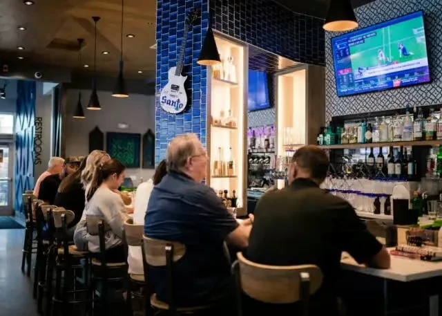 Modern bar interior with patrons, stocked counter, sports TV, Santo guitar.